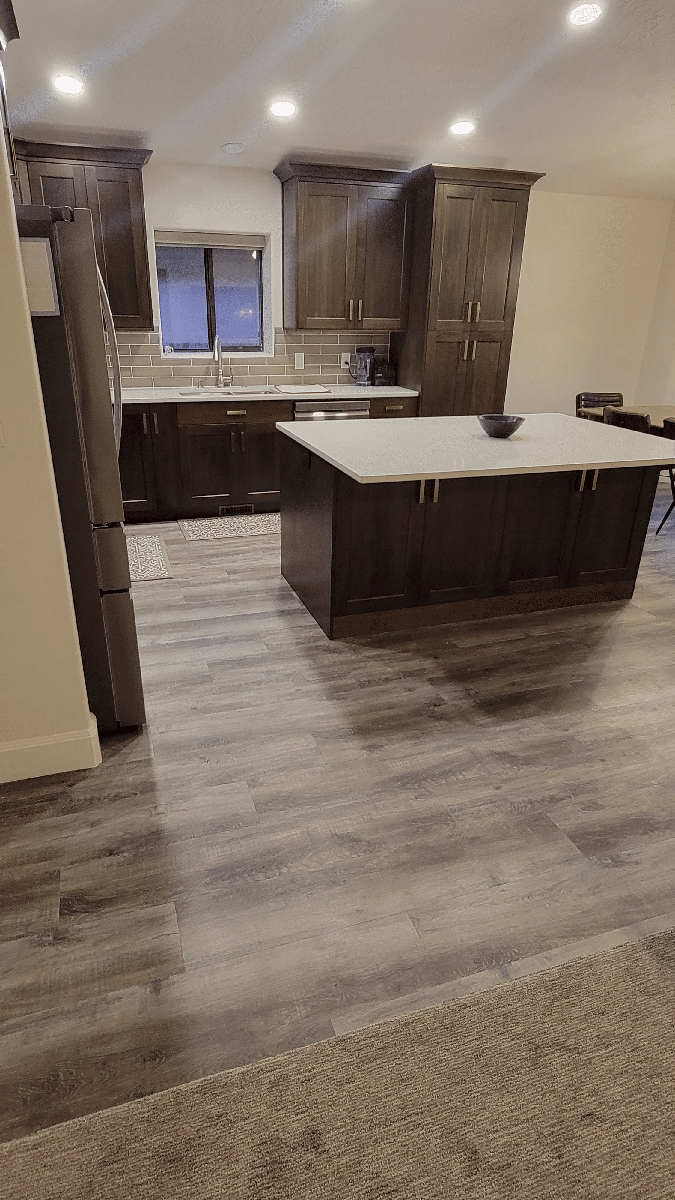 Modern kitchen with dark wood cabinets, central island, stainless steel fridge, and tiled backsplash. Laminate flooring and beige walls.