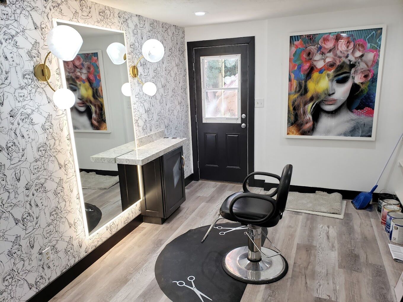 Hair salon interior with a single styling chair, a large mirror, and wall art depicting a woman's face with flowers. Walls have black and white illustrations; broom and dustpan in the corner.