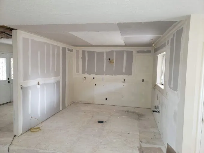 An unfinished kitchen space with drywall, missing appliances, and no flooring. Electrical wires and socket openings are visible on the walls.