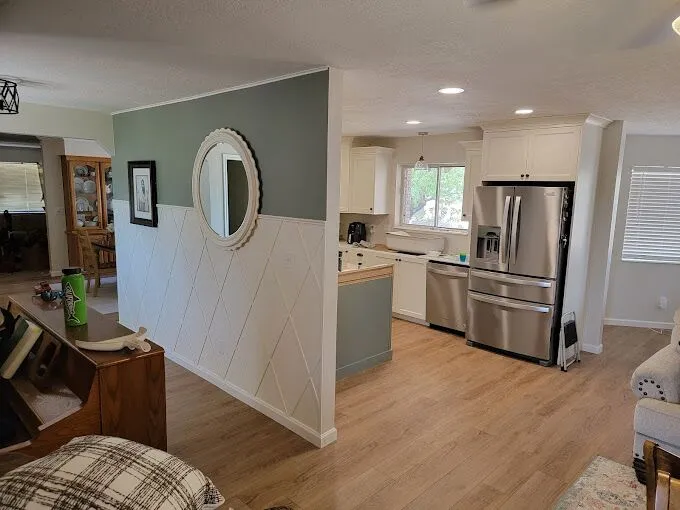 Open-plan kitchen and living area with light wood flooring. Stainless steel fridge, white cabinets, and round wall mirror are visible. Natural light from windows.