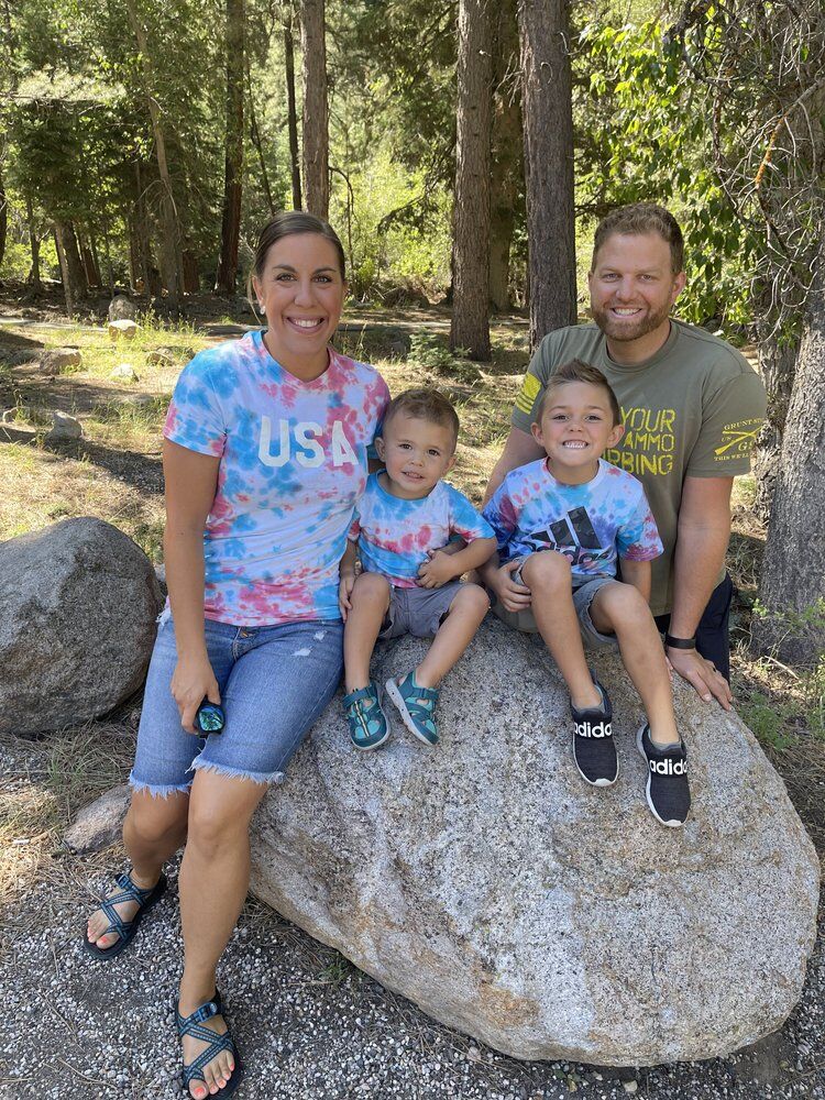About Us: A family of four poses outdoors on a large rock, exuding joy and togetherness. Two adults and two children, wearing vibrant tie-dye shirts, smile brightly against the backdrop of a lush forested area.