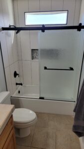 Bathroom with a white bathtub and frosted glass sliding door, white tiled walls, a small window, and black fixtures. A toilet and wooden cabinet are adjacent. A gray towel hangs nearby.