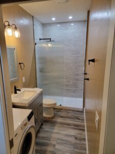 Modern bathroom with wood flooring, featuring a shower with textured glass door, a toilet, a sink with marble countertop, vanity mirror, washing machine, and wall-mounted lights.