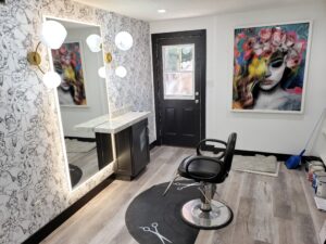 Hair salon interior with a black styling chair, illuminated mirror, artistic wallpaper, a decorative painting, and a broom on the floor next to a black door.