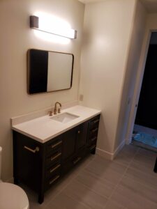 Bathroom with a dark vanity, white countertop, and under-mount sink. A rectangular mirror and a light fixture are above. Gray tiled floor and off-white walls are visible.
