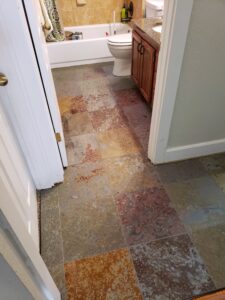 Bathroom with multicolored slate tile flooring, a white toilet, a bathtub partially visible, and a wooden cabinet with a counter.
