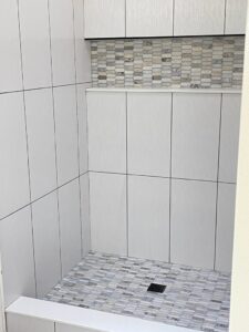 Shower with light gray rectangular tiles, a mosaic tile accent wall and floor, and built-in shelf.