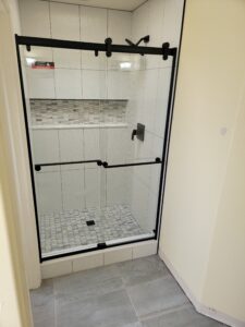 Glass-enclosed shower with black frame, featuring white tiles and a mosaic tile accent strip, located in a tiled bathroom corner.