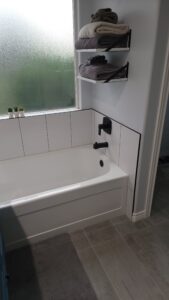 Bathroom with a bathtub, black faucet, and white tiled walls. Two shelves above hold folded towels. A frosted window is to the left, with small plants on the windowsill. Light gray flooring.