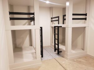 Four empty bunk beds with black ladders and railings in a small room with beige walls and speckled flooring.
