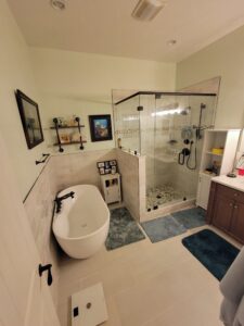 Bathroom with a freestanding bathtub, walk-in glass shower, and wooden vanity. Decor includes wall art and shelving. Blue and gray mats are on the tiled floor.