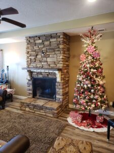 Room with a stone fireplace and a decorated Christmas tree with red and white ornaments. A rug covers the wooden floor, and a lamp is visible to the left.