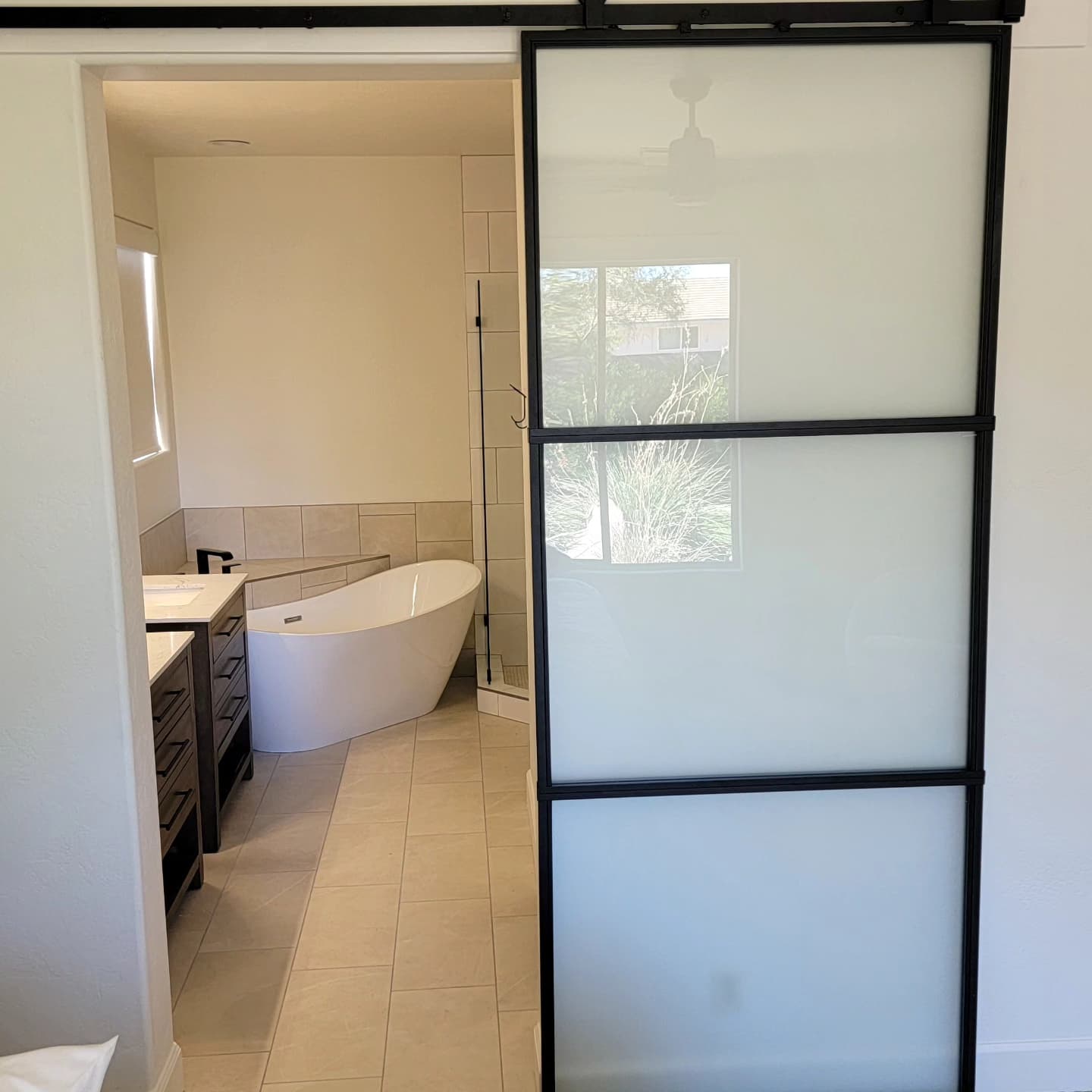 Sliding glass door opens to a modern bathroom with a white freestanding bathtub, beige tile flooring, and a black double-sink vanity.