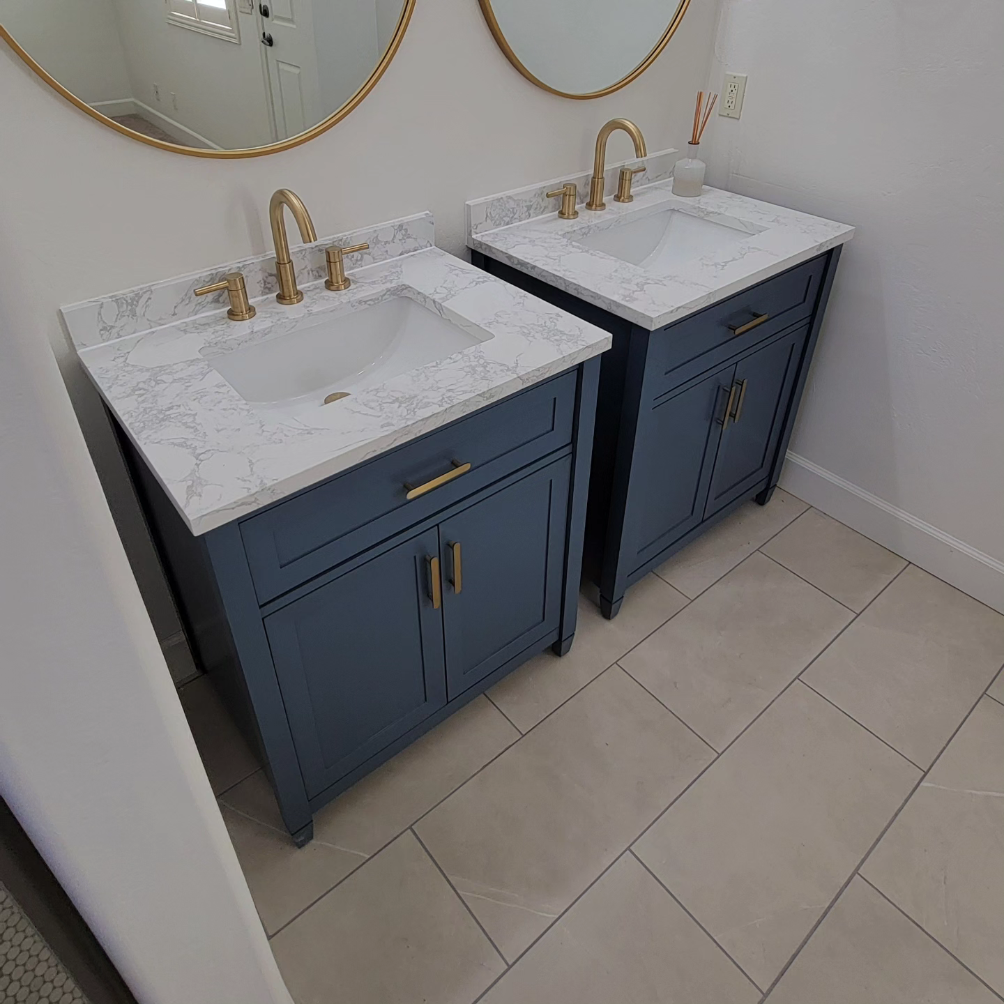 Two blue bathroom vanities with marble countertops, gold faucets, and circular mirrors above, set against a white wall.
