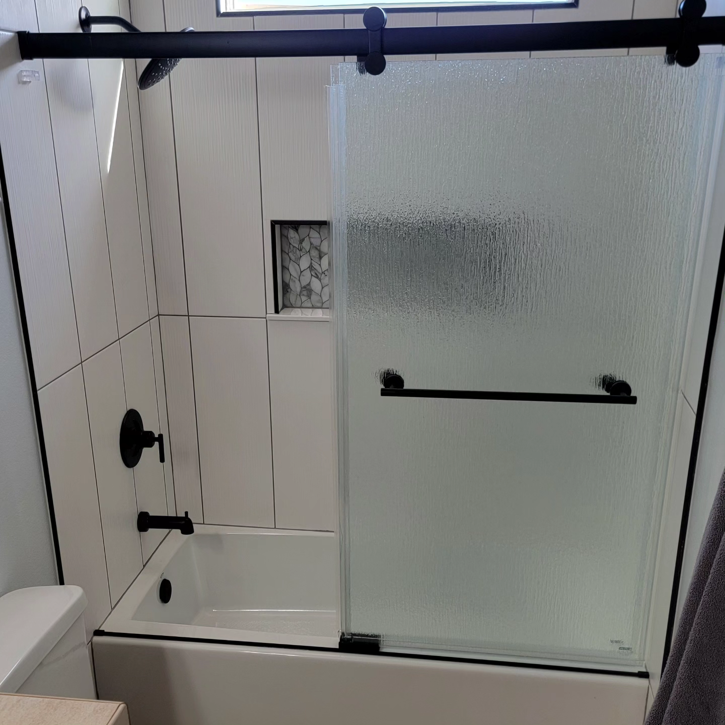Bathroom with a bathtub featuring frosted sliding glass doors, black fixtures, and a tiled wall with a small recessed shelf.