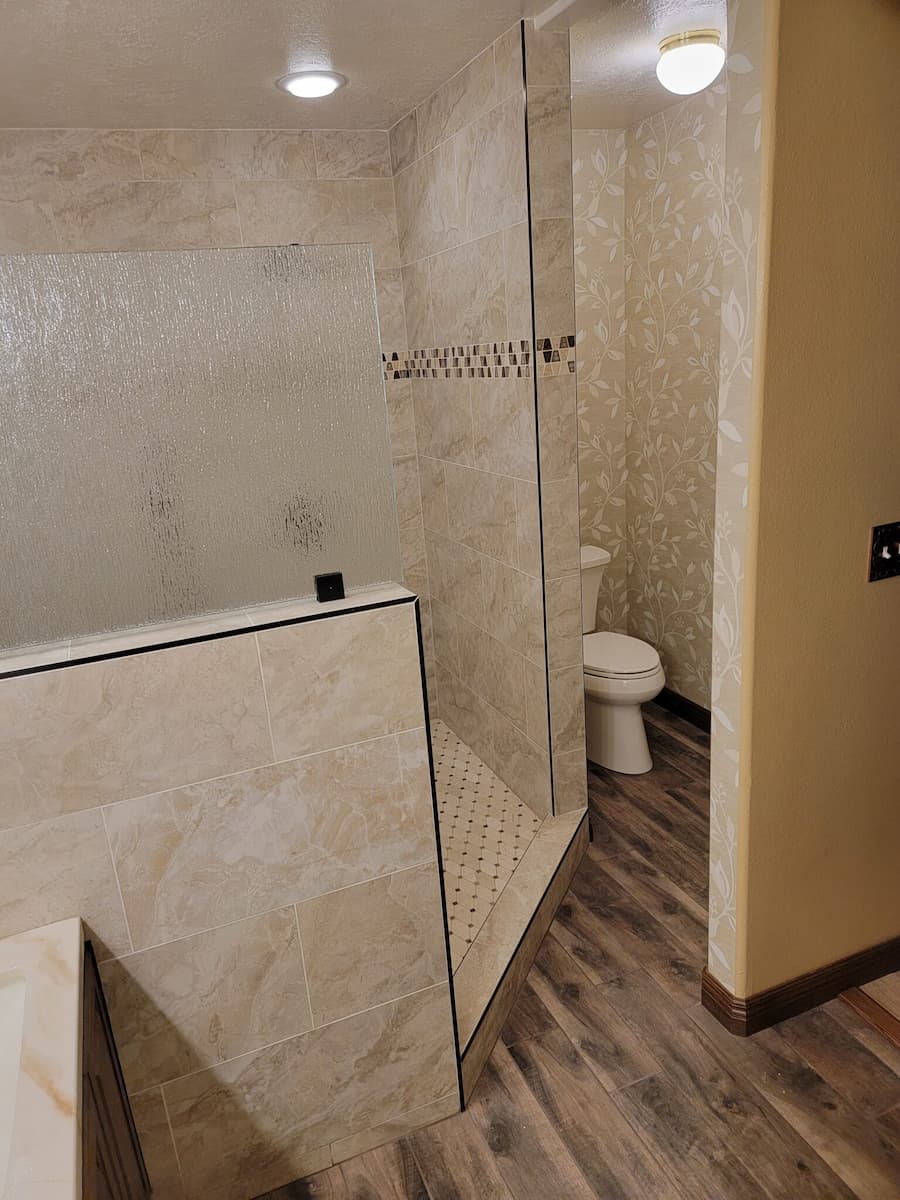 Tile bathroom featuring a walk-in shower with glass partition and geometric pattern, expertly crafted by bathroom remodeler services. Toilet visible in the background. Wood-patterned floor adds contrast to the light walls, creating a harmonious and stylish space.