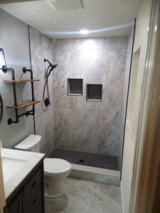 Modern bathroom with a shower area featuring gray marble-like tiles, black fixtures, and built-in shelves. The space includes a vanity, toilet, and wall-mounted shelves.