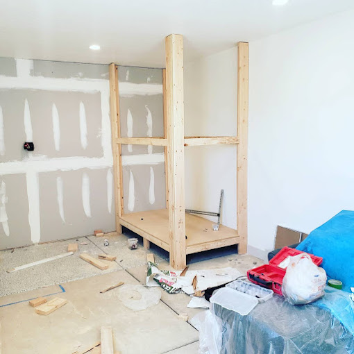 Room under renovation with wooden frame structure, unfinished drywall, scattered tools, and construction materials on the floor.
