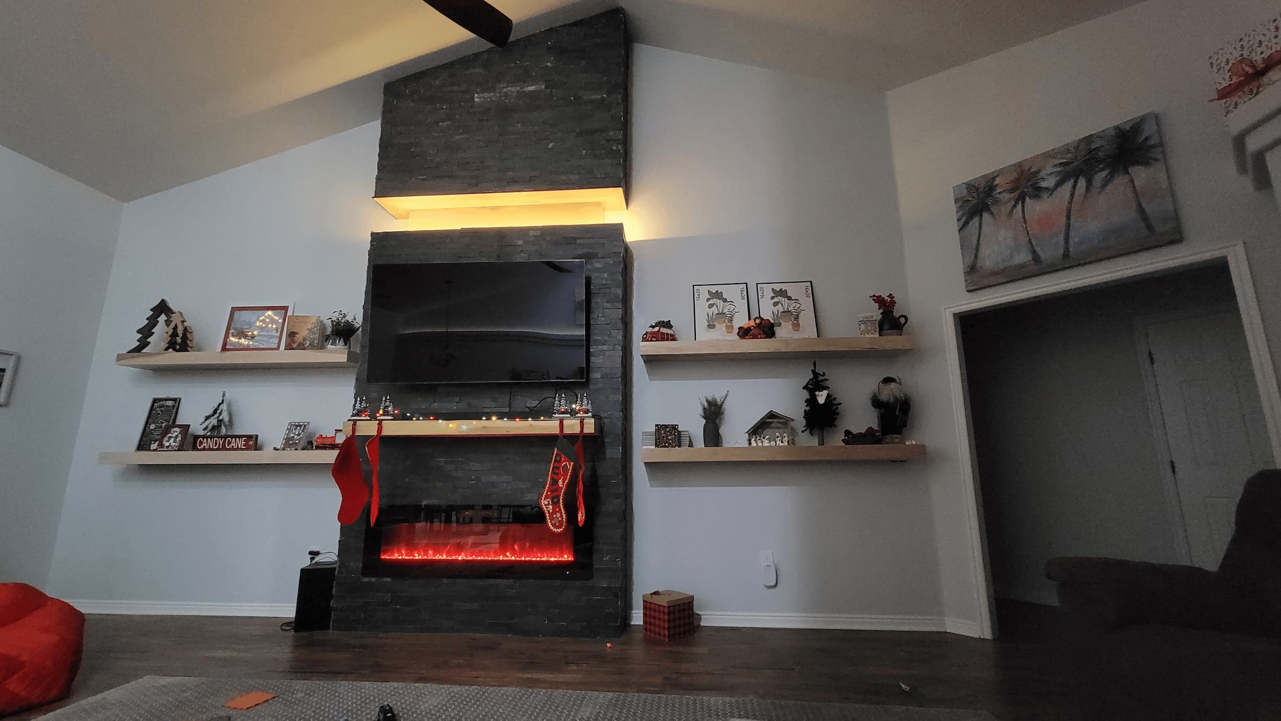 Living room with a dark stone fireplace, mounted TV, and electric fire. Shelves display decorations, including Christmas stockings and festive ornaments. Ceiling fan above, armchair to the right.